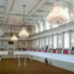 Inside an ornate hall with two tier balconies and chandeliers. Horses walk around on the central floor.