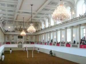 Inside an ornate hall with two tier balconies and chandeliers. Horses walk around on the central floor.