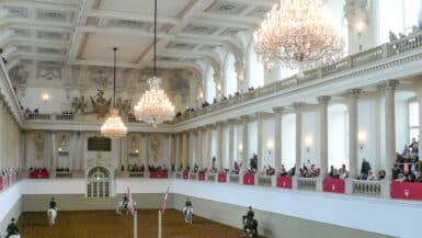 Inside an ornate hall with two tier balconies and chandeliers. Horses walk around on the central floor.