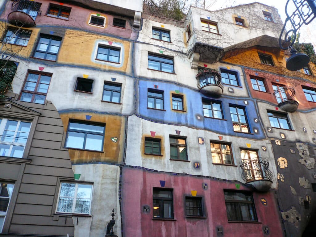Looking up at a tall building which is painted in different patches - white, blue, pink. Trees on rood and on walls. Iron balconies on some flats.