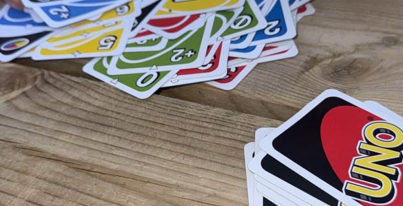 Someone is dealing cards on a wooden table. Their hands are picking up a car from a loose pile on the table. An organised pile to the right shows the cards are UNO