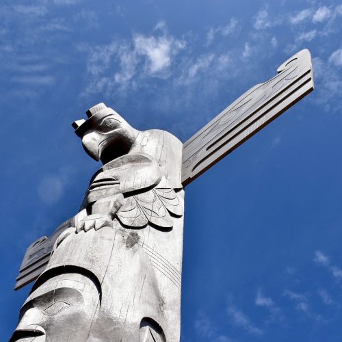 Blue sky with patchy white cloud. A carved grey totem rises up from the bottom left of the frame into the middle of the photo - it as a man's face at the bottom and a thunderbird / eagle at the top with open wings
