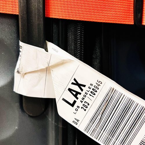 A white luggage label with the words LAX Los Angels on it along with the numbers in black and a bar code. The label is attached to the black handle of a dark suitcase. You can just see a woven orange strap around the top of the suitcase