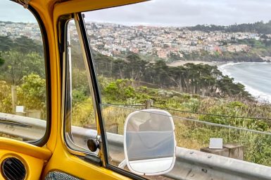 View through window of yellow can across a bay with trees in foreground, houses on the cliff side and water to the right. Grey sky above