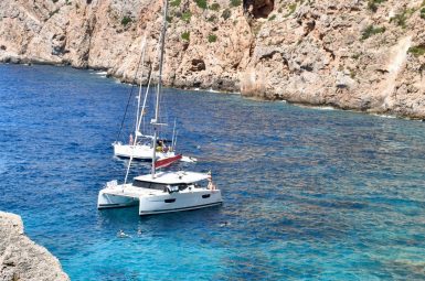 Rocky cliff in background and to the bottom left of photo. A body of turquoise water fulls the rest of the frame and two white boats sit on the water. One is a small sail boat (sails not up) in background and front boat is a small catarmaran