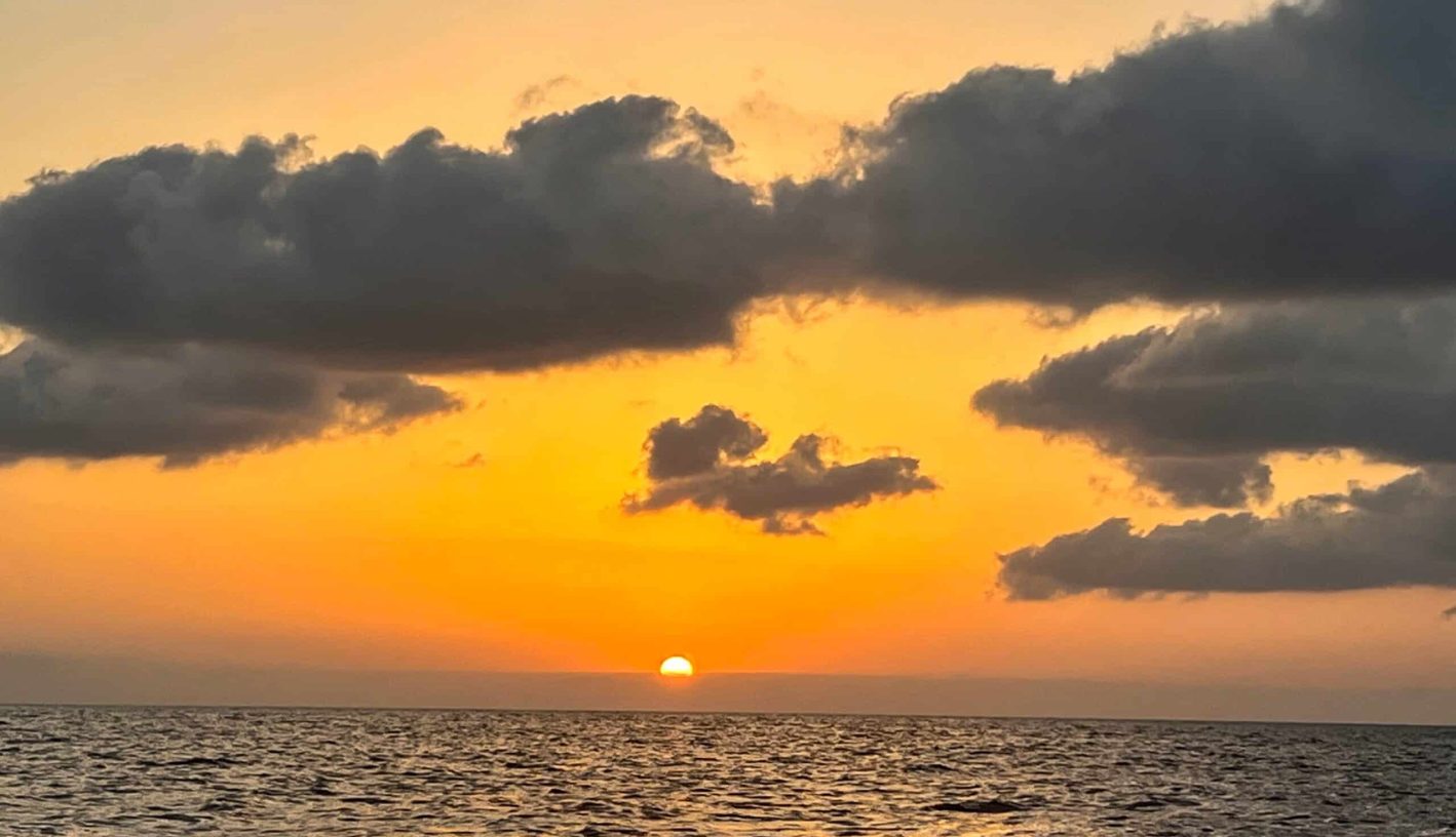Sunset photo - bottom third is a dark body of sea with the golden glow of the sun across the water. The sun is settling on the horizon. Sky is deep orange near the middle, getting paler peach colour to the top. Big fluffy grey clouds across the top of the photo
