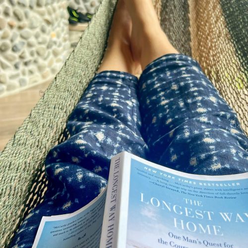 Photo taken from inside a green and yellow woven hammock. You can see the legs of the person in hammock. they are wearing blue and white trousers. There is a book open with the title The Longest Way Home. Stone wall in background