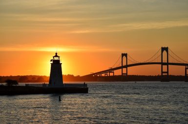 View across a harbour at sunset with a two span suspension bridge silhouetted on the horizon. Into the left is a lighthouse in silhouette with the sunset directly behind it.