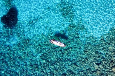 Drone shot of a paddle board floating on a turquoise blue sea. The water is so pure you can see the rocks underneath
