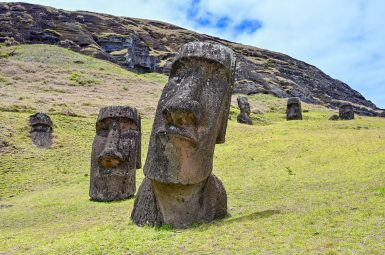 Looking up a grassy bank. A large carved stone statue rises up from the grass - human head. A smaller one to the left and two in the distance to the right. Rocks and cloudy sky above