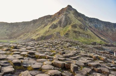 A large causeway of connected hexagonal stones leads from the front of the frame towards the centre middle. Behind is a green rocky cliff rising to a point in the centre. Grey sky behind.