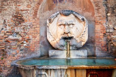 Face carved from stone sits above a bath of turquoise water in front of a red brick wall. It is an old water fountain