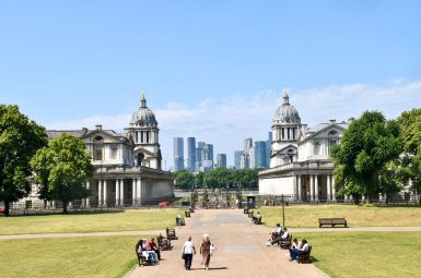 view from an elevated position looking down a broad path with lawns on either side. Benches on the path with people sitting on the benches and walking down the path. There are wrought iron railings at the end of the lawn with a red double decker bus passing. Behind the bus are modern office tower blocks in the back ground and old fashioned white buildings with columns between the bus and the office blocks
