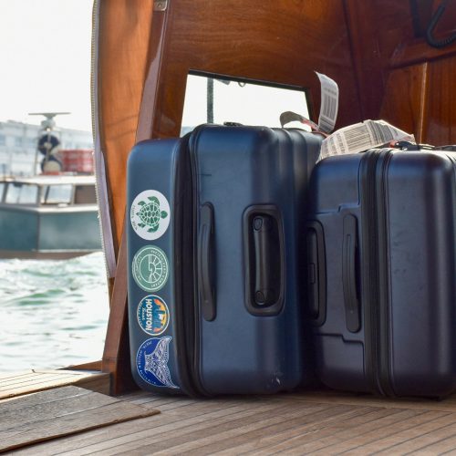Two navy blue suitcases lie on their sides on the floor of a wooden boat. The left suitcase is slightly bigger and features 4 stickers (one says Houston on it). The right hand suitcase has an orange strap. Both cases have their white airline tags showing on the top. To the left hand side of the boat, you can see the water through the side and a boat sailing past
