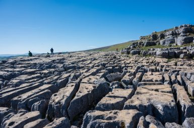 View along a platform or pavement of rock which is broken up with ridges in between the rocks as it created through glaciation. A couple of people are walking on the rocks to the left hand side. Blue sky above. Rocks to the right