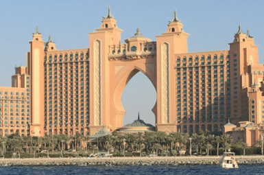 Arabic style architecture on tall pink building which could be a hotel. Row of palm trees in front and sea in foreground. Pale blue sky behind