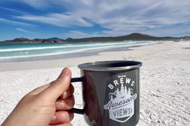 A hand in the foreground holds out a black metal cup which says 'Warm Brews and Awesome Views!'. Behind is a sweeping powder white beach and turquoise waves across the bay. On the horizon are some hills with a cloudy blue sky above.