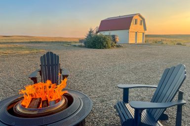Fire pit and green chair in the foreground. White barn with red roof in background against a setting sun