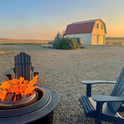 Fire pit and green chair in the foreground. White barn with red roof in background against a setting sun