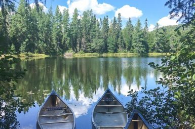 View through the trees to a still lake with fir trees on the opposite right. Blue sky with white fluffy clouds. The sky is mirrored in the flat waters of the lake. Three canoes are lined up on the front bank of the lake.